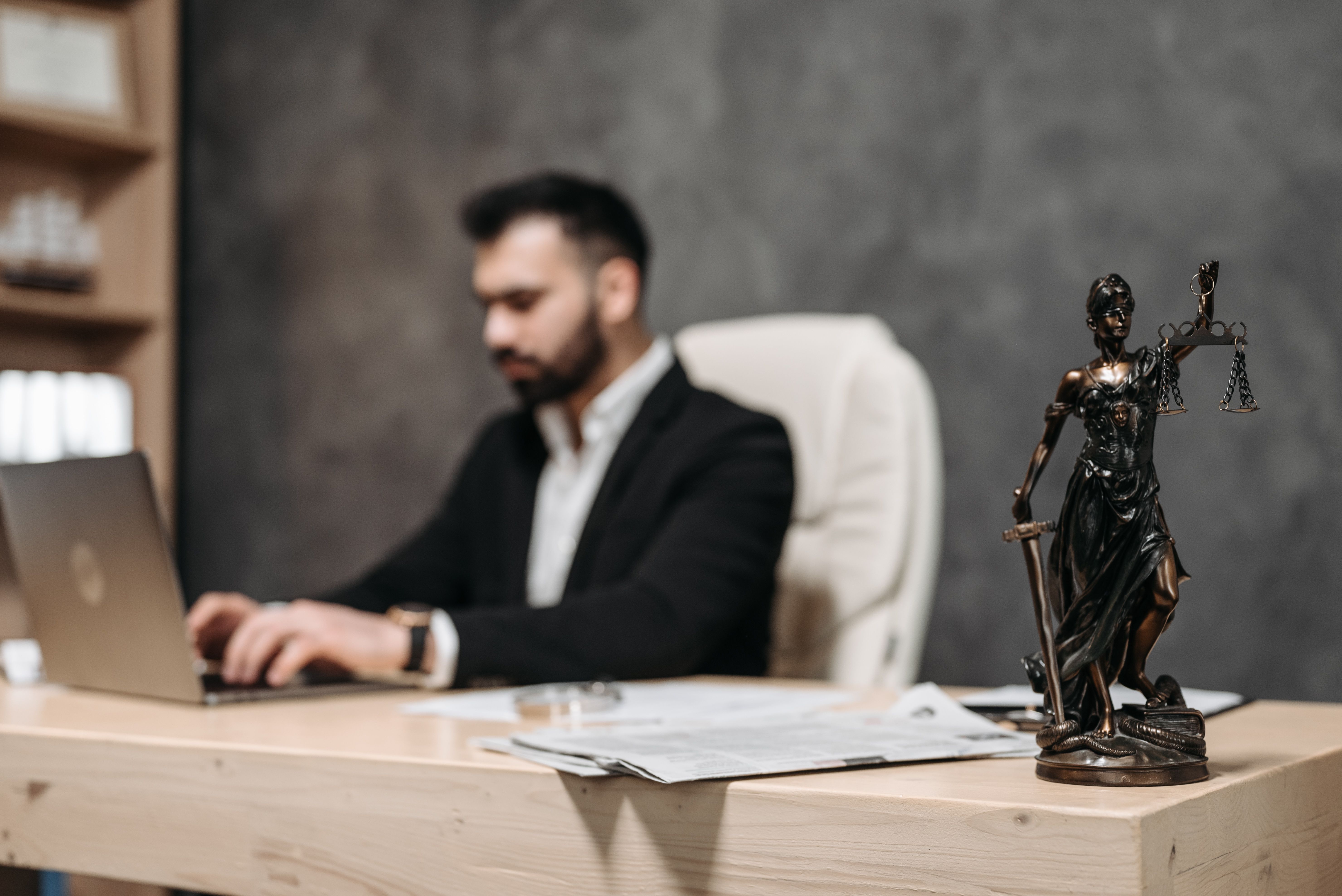 Businessman working on a laptop in an office with a Lady Justice statue on the desk, symbolizing law firm finances
